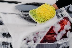 Back view of mans hand with sponge washing car hood and lights. Black auto covered with foam. Close up macro. Service station photo