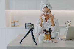 Pretty happy female blogger records streaming video for her blog gives tutorial how to apply beauty patches gives advice about skin care dressed in bathrobe looks at camera poses at table in kitchen photo