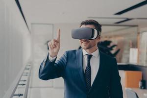 Smiling bearded office employee in suit wearing in VR glasses working in augmented reality in office photo