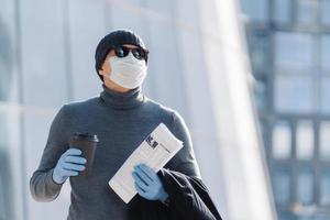 Horizontal shot of young man in medical mask prevents infectious pneumonia, coronavirus pandemic, virus epidemic, drinks coffee to go, going to work, poses outside, has thoughtful expression photo