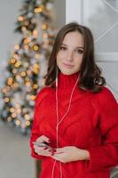 tranquila mujer encantadora y despreocupada con piel sana, vestida con suéter rojo, usa teléfono móvil y auriculares, disfruta del ambiente doméstico, pasa la Navidad en casa. concepto de personas y tecnología foto