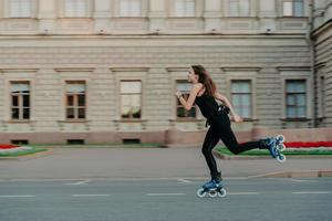 Outdoor physical activities. Healthy young woman has slim fit figure leads active lifestyle spends free time outside rides on rollerblades poses alone in open air against building background photo