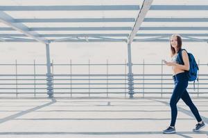 Positive slim fitness girl going on training, being photographed in motions, carries bag, holds modern cell phone, uses up to date earphones, stands outdoor, copy space aside for your promotion photo