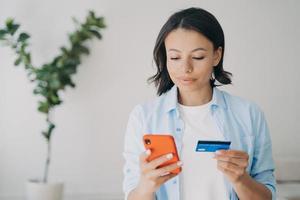 Female holding bank credit card, smartphone, using online banking services paying purchase at home