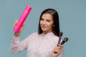 Women and beauty concept. Dark haired positive woman holds hairspray and two cosmetic brushes, prepares for date, cares about her appearance, wears elegant shirt, isolated on blue background photo