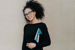 Positive curly African American student carries notepad and diary, wears black clothes, transparent glasses, enjoys studying in university, isolated on grey background. Cute woman with notebook photo