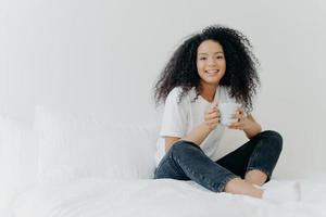 Young pretty African American woman rests in bed at home, drinks hot tea in morning, enjoys domestic atmosphere, tasty drink while relaxes in bedroom. People, leisure, rest and lifestyle concept photo