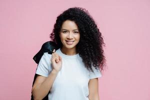 Portrait of positive ethnic woman wears white t shirt, carries jacket on shoulder, has cheerful expression, ready for picnic, enjoys day off, poses indoor against rosy background. People, lifestyle photo