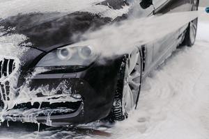 lavado automático en lavado manual de autos. vehículo negro cubierto de agua y espuma de lavado limpiada por un chorro de agua a alta presión foto