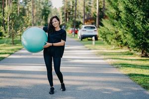 Full length shot of active sporty woman holds big fitness ball exercises outdoor walks on road near forest leads healthy lifestyle stays fit dressed in sportswear. People gymnastics and aerobics photo