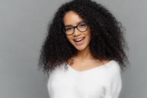 Indoor shot of happy young lady has Afro hairstyle, smiles broadly, glad to be promoted, wears optical glasses and white sweater, models over grey background. Ethnicity, emotions, pleasure concept photo