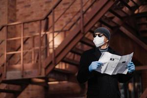 Horiontal shot of pensive man poses with newspaper, wears protective mask and rubber gloves, looks aside, thinks about self isolation, quarantine and coronavirus pandemic. People and covid 19 photo