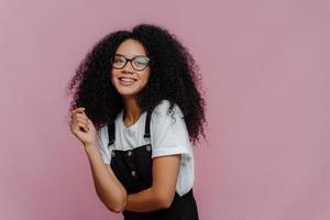 Studio shot of happy dark skinned lady with crisp hair, has dreamy expression, wears optical glasses, stylish clothes, imagines something pleasant, stands against bright violet background, free space photo