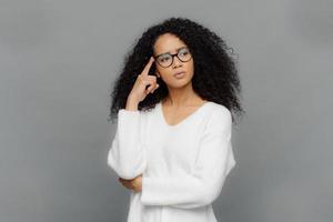 Troubled serious female keeps fore finger on temple, looks aside, has curly hair, being deep in thoughts, wears white jumper and optical glasses, isolated on grey background. People and thinking photo