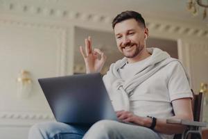 Modern bearded guy makes okay gesture looks gladfully at laptop display wears earbuds has video call wears t shirt jeans jumper tied over shoulders poses in modern apartment. Technology concept photo