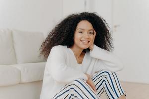 Photo of relaxed delighted African American woman with curly hairstyle, smiles gently at camera, wears white sweater and striped trousers, sits on floor near couch in living room of modern apartment