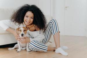 Indoor shot of pleased curly haired female model poses for photo with jack russell terrier dog, embraces pet tenderly, sits on floor with beloved animal near comfortable sofa, spend time together