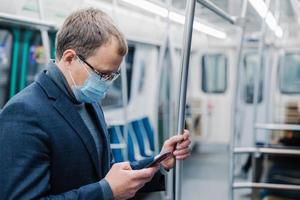 Serious man commutes to work in underground wears virus protective mask being always in touch with modern cellular poses in empty subway or metro. Distance concept. Public transport during coronavirus