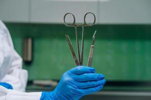 A rubber gloved hand holds two scalpels and a clamp photo