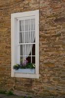 A white-framed window with a flower box set into a brown stone wall. photo