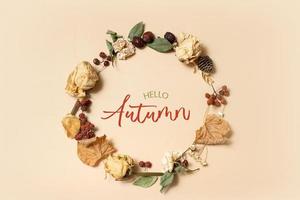 Autumn composition with Hello Autumn text. Wreath made of dried leaves and flowers on pastel beige background. Flat lay, top view photo