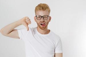 Dislike sign, hand gesture. Young man showing thumb down. Unhappy and sad guy with dissapoint face in glasses and blank template white t shirt with copy space. Mockup t-shirt. Disgust emotions photo