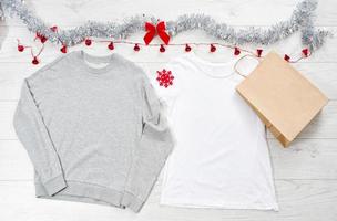 cierre la camiseta blanca con plantilla en blanco, sudadera con capucha con espacio para copiar. concepto de vacaciones de navidad. camiseta de bolsa de papel de maqueta de vista superior, lazo rojo sobre fondo de madera blanca. feliz año nuevo decoraciones foto