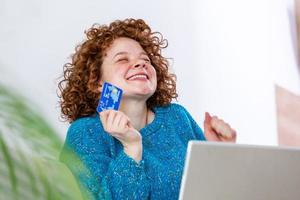 Happy Young Woman On Sofa Shopping Online With Debit Card. Beautiful girl using laptop computer for online shopping at home exited for finding item on sale photo