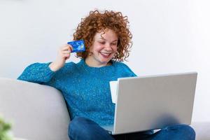 Happy Young Woman On Sofa Shopping Online With Debit Card. Beautiful girl using laptop computer for online shopping at home exited for finding item on sale photo