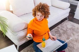 Cute female red hair freelancer reading text messages while sitting on floor with open computer in modern home, young creative woman work on laptop while having orange juice photo