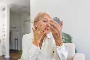 Image of feminine mature woman with short blond hair holding mirror Senior woman applying anti-aging lotion photo