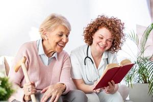 mujer cuidadora leyendo un libro mientras se sienta con una anciana feliz en un hogar de ancianos. feliz anciana sentada en un sofá blanco y escuchando a la enfermera leyendo un libro en voz alta foto