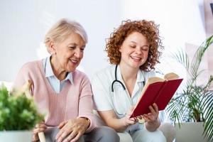 Woman caregiver reading a book while sitting with happy senior woman at nursing home. Happy elder woman sitting on white sofa and listening to nurse reading a book out loud photo