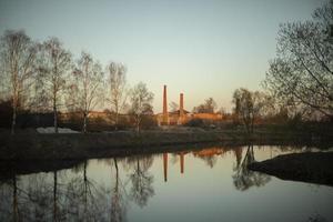 View of factory. Brick pipes of plant. Landscape with lake and birch trees. photo