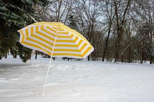 A bright yellow-striped beach umbrella stands on white snow in a park on a snowy glade photo