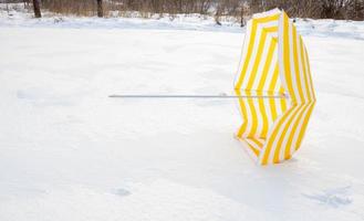 Fallen bright yellow striped beach umbrella on white snow in a park in a snow-covered clearing photo