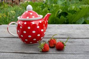 Red vintage teapot with a polka dot pattern on a wooden background next to strawberries. Place for text photo