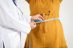 una doctora con uniforme blanco sostiene una cinta métrica alrededor de la cintura de una mujer obesa con un vestido amarillo que tiene problemas de salud y obesidad. concepto de tratamiento del paciente, pérdida de peso. foto