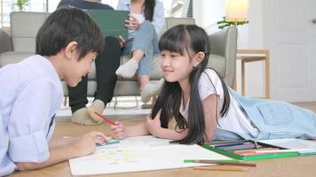 Asian Thai siblings are lying on living room floor, drawing homework with colored pencils together, parents leisurely relax on a sofa, lovely happy weekend activity, and domestic wellbeing lifestyle. video