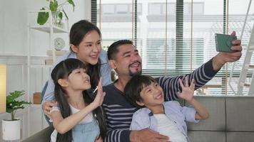 Happy Asian Thai parents, and children online video call with family via internet on sofa in home living room, lovely weekend, and domestic wellbeing lifestyle with internet technology mobile phone.