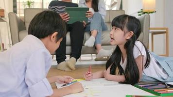 Asian Thai siblings are lying on living room floor, drawing homework with colored pencils together, parents leisurely relax on a sofa, lovely happy weekend activity, and domestic wellbeing lifestyle. video