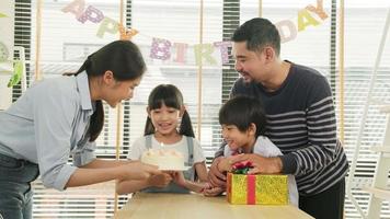 gelukkig Aziatisch Thais familie, jong kinderen verrast door verjaardag taart, geschenk, blazen uit een kaars, en vieren partij met ouders en broers en zussen samen Bij dining tafel, welzijn huiselijk huis speciaal evenement. video