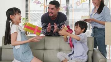 gelukkig Aziatisch Thais familie, jong kinderen verrast door verjaardag taart, geschenk, blazen uit kaars, en vieren partij met ouders en broers en zussen samen in leven kamer, welzijn huiselijk huis evenement levensstijl. video