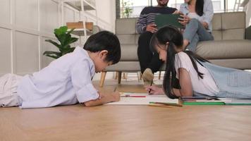 les frères et sœurs thaïlandais asiatiques sont allongés sur le sol du salon, dessinant ensemble des devoirs avec des crayons de couleur, les parents se détendent tranquillement sur un canapé, une belle activité de week-end heureux et un style de vie de bien-être domestique. video
