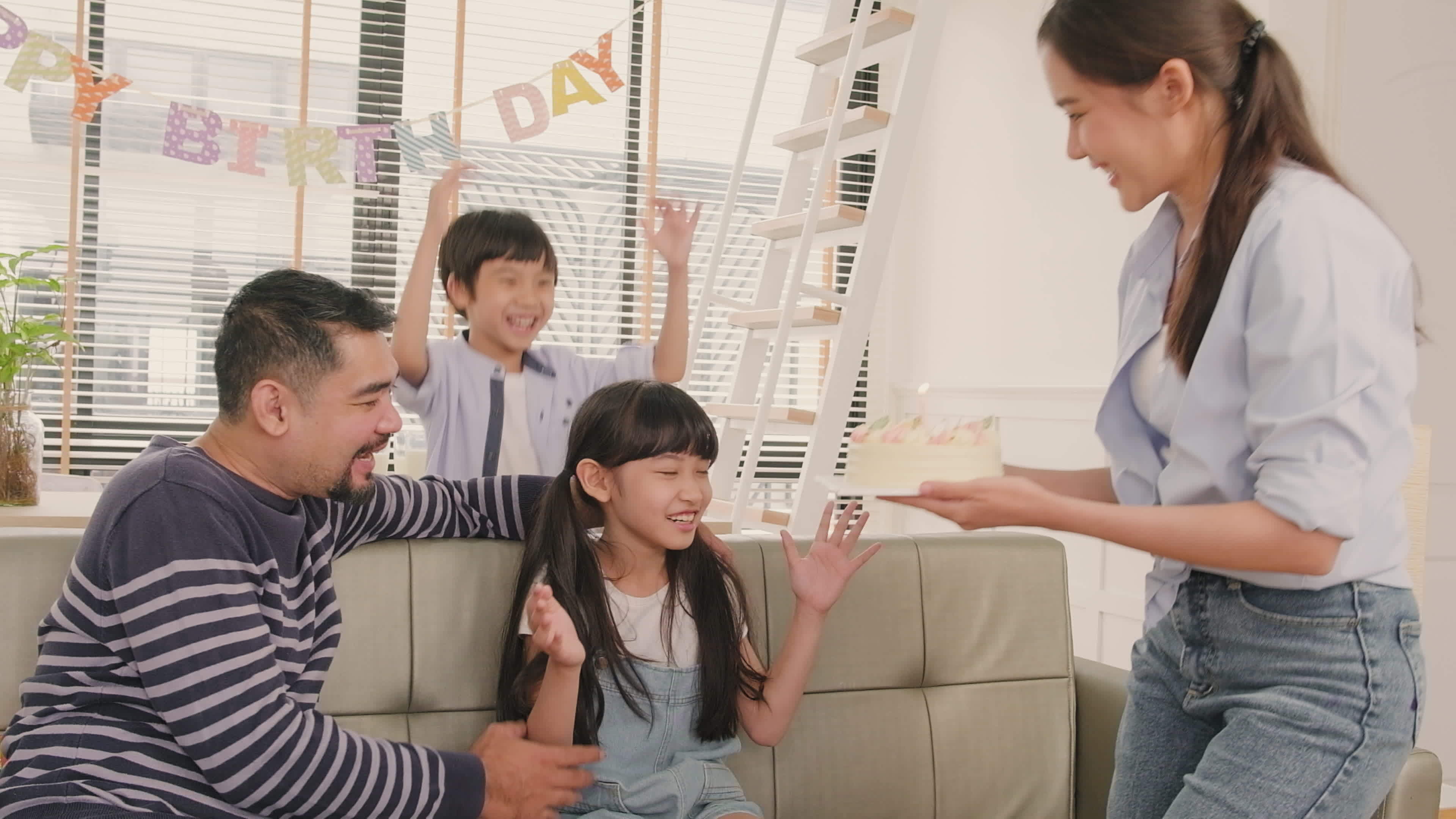 Happy Asian Thai family, young daughter is surprised with birthday cake, blows out candle, and cheerful celebrates party with parents together in living room, well-being domestic home event lifestyle.