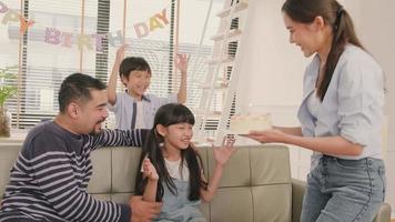 feliz família tailandesa asiática, filha jovem é surpreendida com bolo de aniversário, apaga vela e alegre celebra festa com os pais juntos na sala de estar, estilo de vida de eventos domésticos domésticos de bem-estar. video