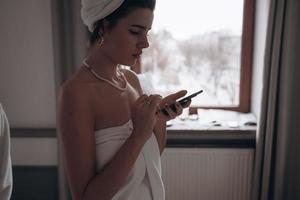 mujer de belleza en bata de baño y con una toalla en la cabeza usando un teléfono inteligente foto