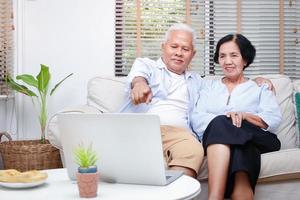 una pareja asiática de edad avanzada ve los medios en línea en su computadora portátil en la sala de estar de su casa. concepto de vida después de la jubilación. la distancia social previene la propagación del coronavirus foto