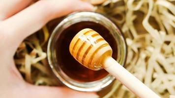 Hand holding honey jar with dipper on wood background with paper shreds. Close up macro shot. Health and beauty product sustainable lifestyle concept. photo