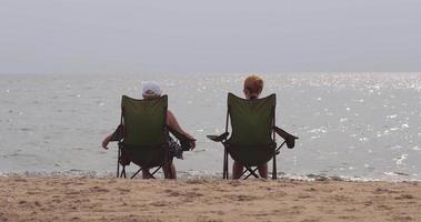 två flickor är Sammanträde i Sol solstolar på de Strand av ett tömma strand nära de vatten video